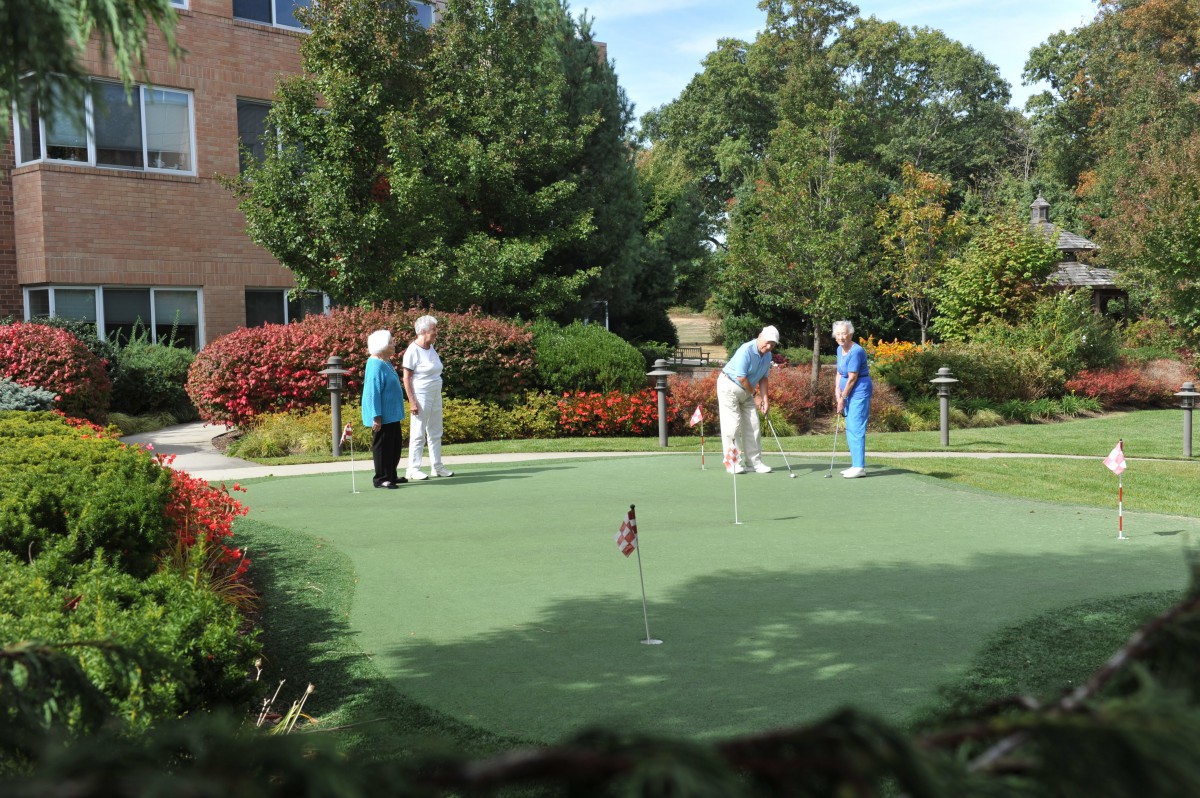 Group of people playing on a putting green outdoors surrounded by trees and gardens.
