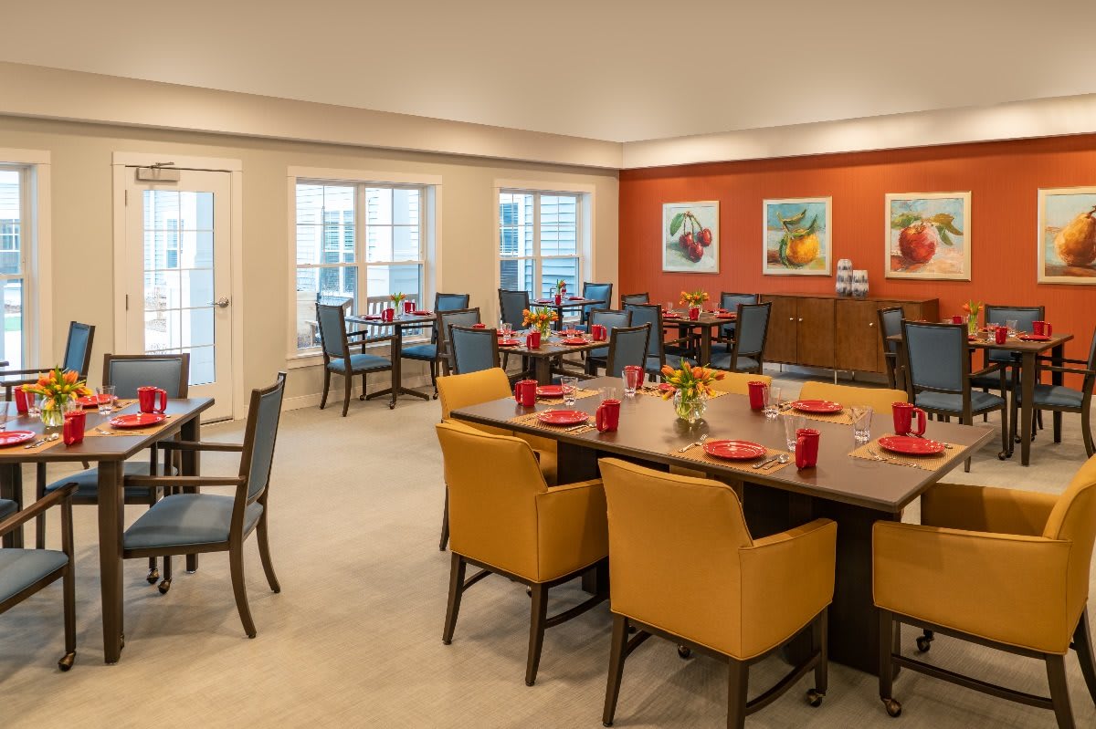 A well-decorated dining area featuring tables with red dishware, surrounded by comfortable chairs and adorned with colorful art on an orange accent wall.