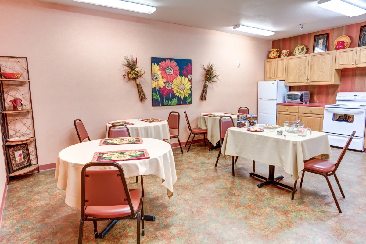 A cozy dining room with arranged tables, decorated walls, and a kitchenette in the background.