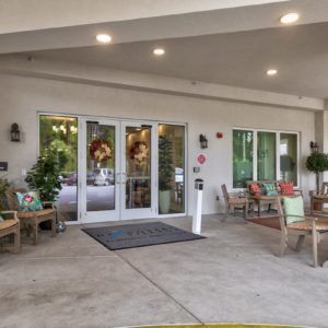 The image shows the entrance of a building with glass doors and outdoor seating, featuring chairs and decor.