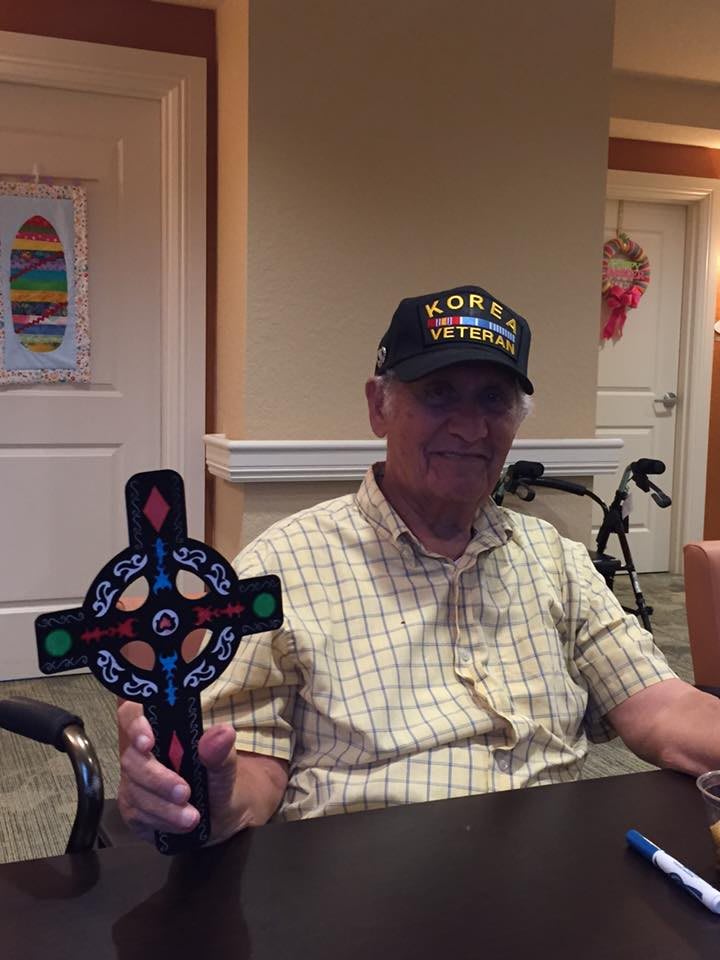 An elderly person wearing a hat and plaid shirt is sitting indoors, holding a decorative cross.