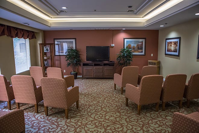 A cozy lounge area with arranged chairs facing a television, featuring decorative elements like plants and paintings.