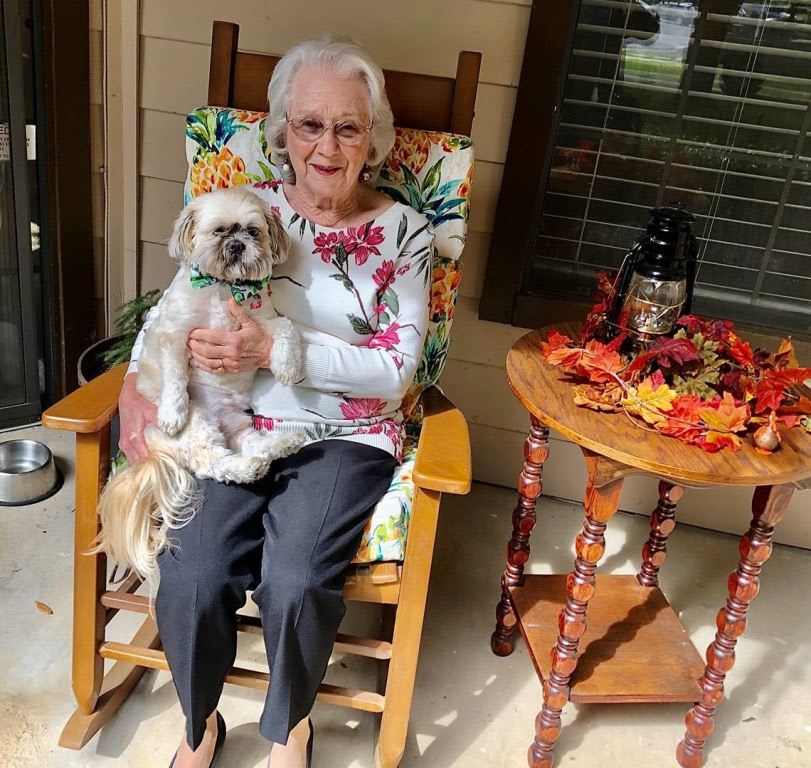 An elderly woman sitting on a porch in a rocking chair, holding a dog with autumn decorations on the side table.