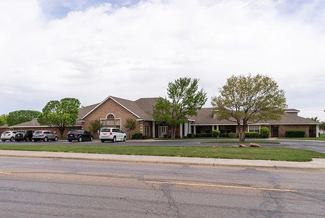 A suburban building with a parking lot, surrounded by trees and cars on a cloudy day.