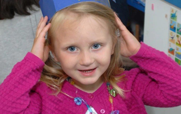 A young girl smiling while adjusting a blue hat.