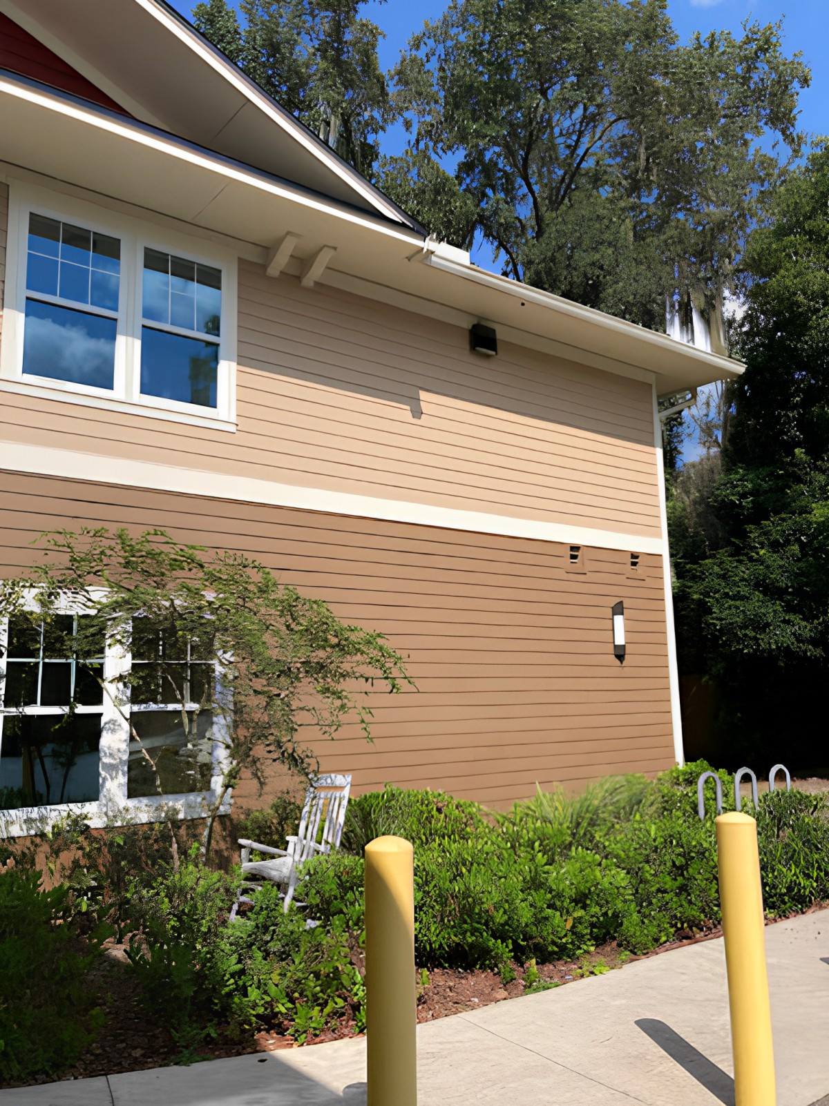 A two-story building with siding exterior, surrounded by greenery and trees on a sunny day.