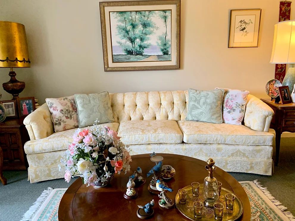 A cozy living room features a plush cream sofa adorned with patterned cushions, a wooden coffee table displaying a floral centerpiece, and elegant wall art.