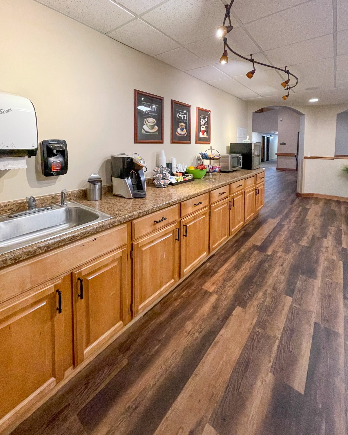 An office kitchenette featuring a long countertop with a sink, coffee machine, and various appliances on a wooden floor.