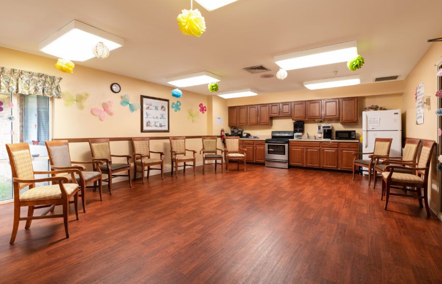 An empty room with wooden flooring, rows of chairs along the wall, and a kitchen area in the background.