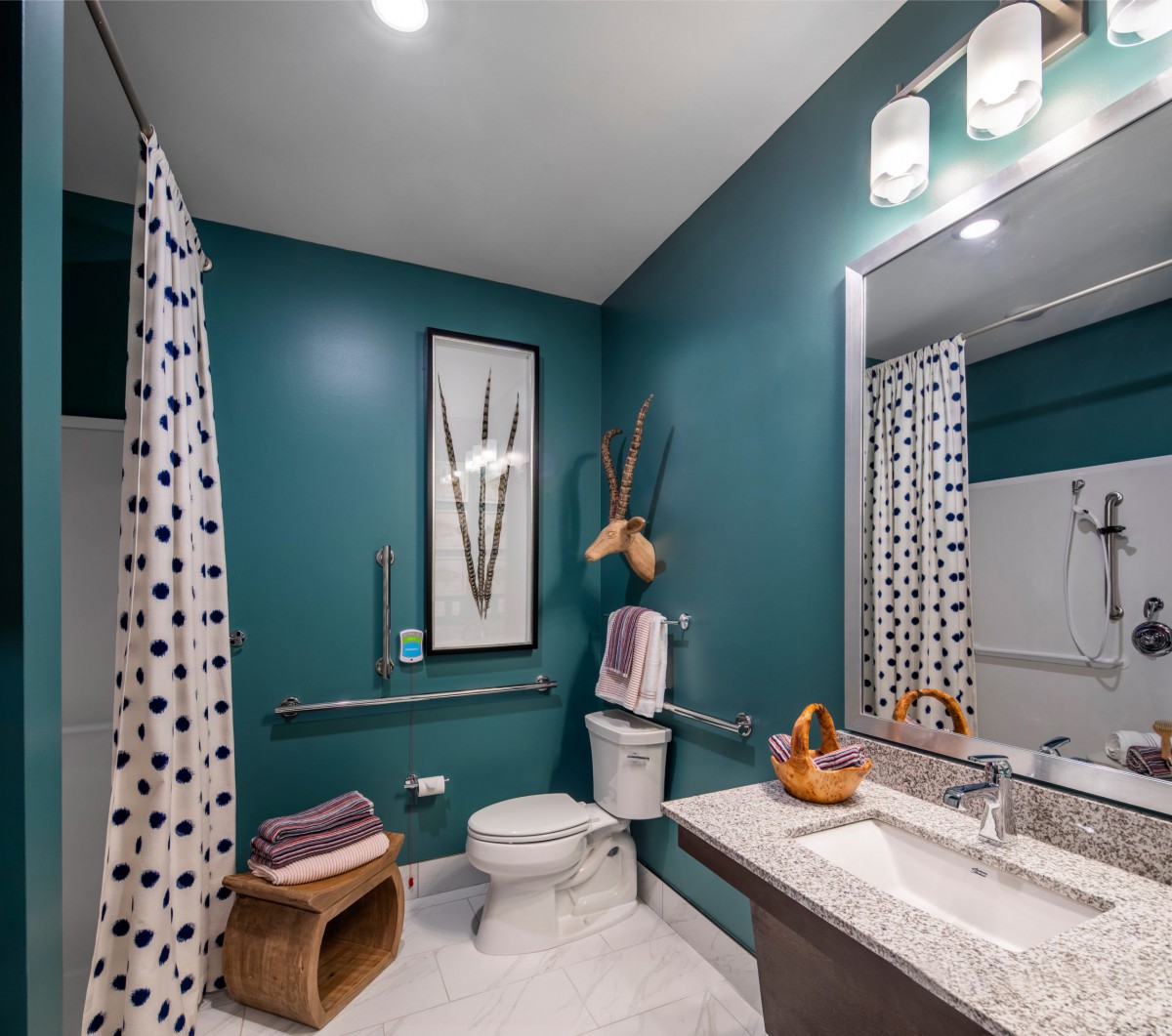 A modern bathroom with teal walls, featuring a white sink and countertop, a shower curtain with polka dots, and decorative artwork.