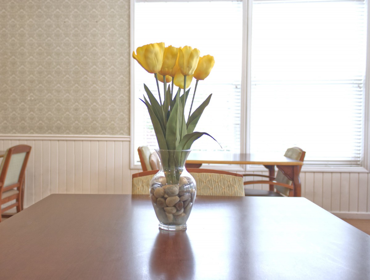 A bouquet of yellow tulips in a decorative vase sits on a wooden table in a well-lit room.