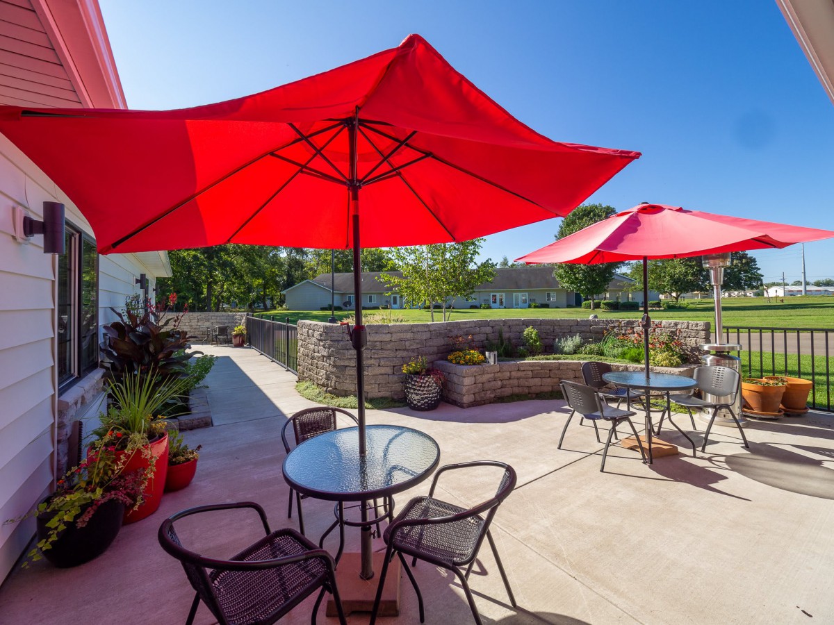 A sunny outdoor patio featuring red umbrellas, tables, and chairs in a garden setting.
