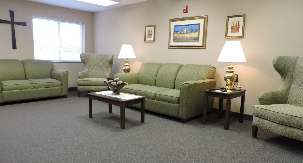 A cozy waiting room with a green couch, armchair, and coffee table adorned with decorative items, illuminated by two lamps and complemented by framed artwork on the walls.