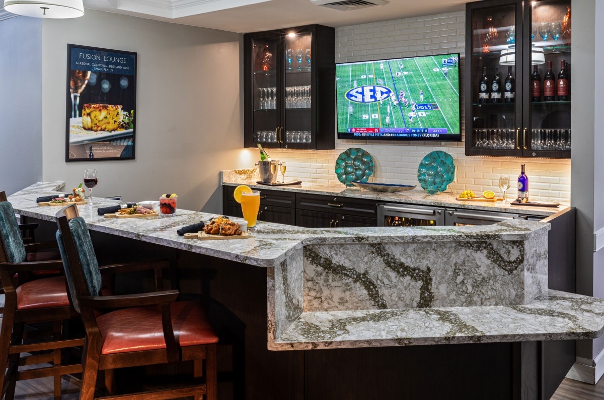 A modern home bar setup featuring a granite countertop with seating, decorated with vibrant accents and a television displaying a football game.