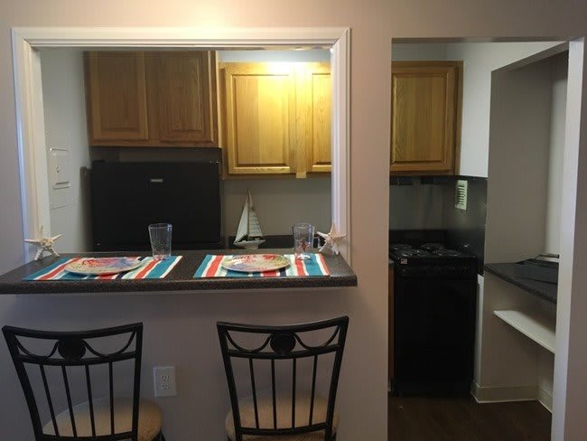 A compact kitchen with a counter and bar stools, featuring wooden cabinets and a small fridge.