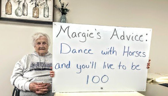 An elderly woman holding a sign with advice: 'Dance with Hope and you'll live to be 100.'