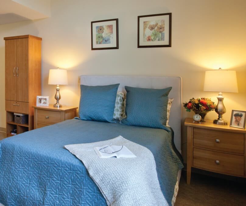 A cozy bedroom with a blue bedspread and matching decorative pillows, flanked by nightstands and lamps, featuring framed artwork on the wall.