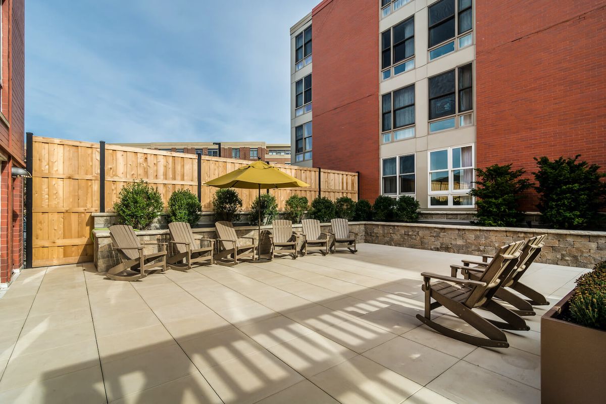 An outdoor patio area with wooden chairs and a yellow umbrella, adjacent to a modern building.