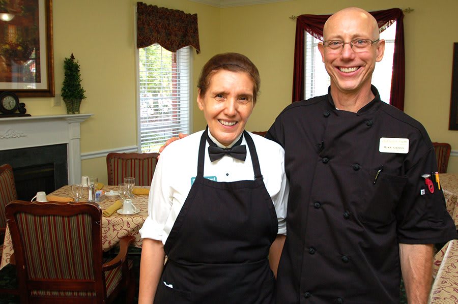 Two restaurant staff members dressed in uniforms and aprons, smiling indoors.