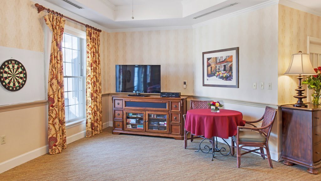 A cozy living room with a TV on a wooden stand, a small red table, and elegant curtains.