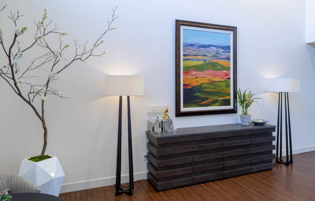 A modern interior featuring a floor lamp, framed landscape painting, and decorative plants on a dark wooden dresser.