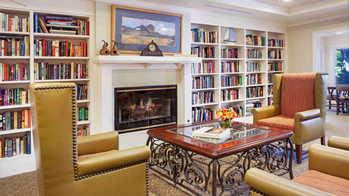 A cozy library room featuring a fireplace, surrounded by bookshelves and comfortable seating around a central table.