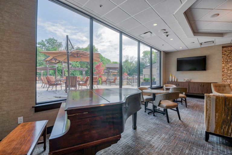 A modern lounge with a grand piano and seating area, featuring large windows overlooking a patio with umbrellas.