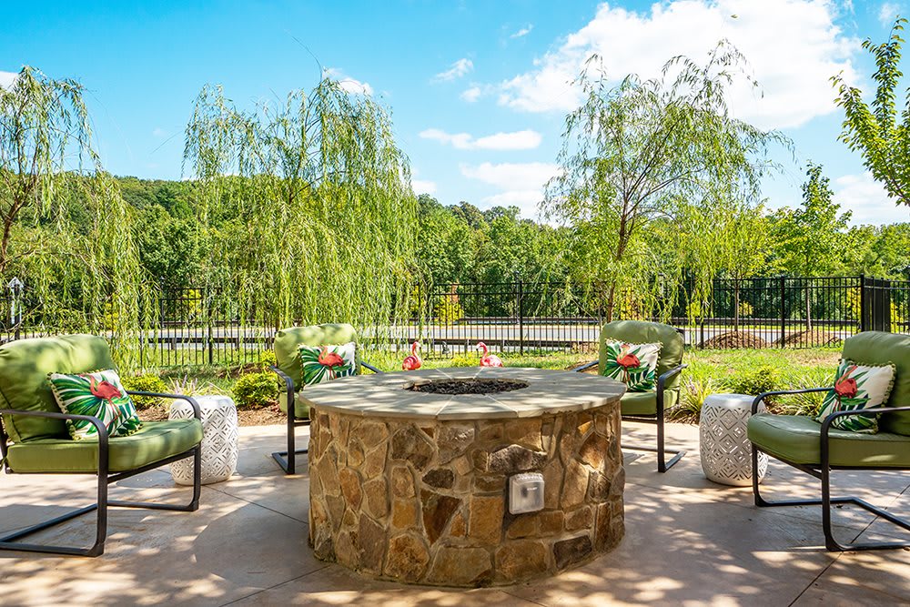 An inviting outdoor patio featuring a stone fire pit surrounded by chairs, set against a backdrop of lush greenery and trees.
