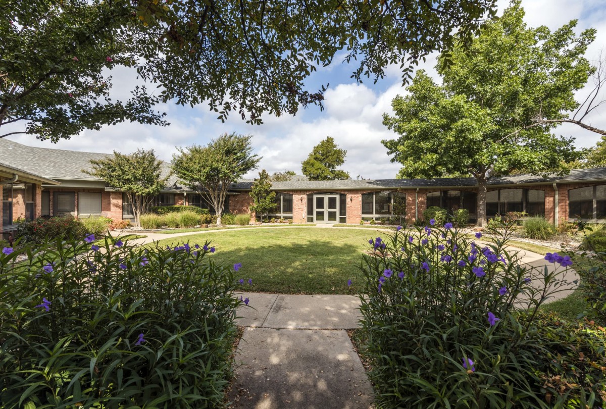 A tranquil courtyard with a pathway leading to a brick building surrounded by lush greenery and trees.