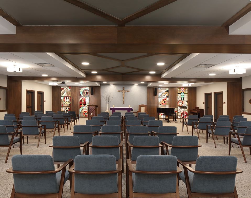 A chapel interior featuring rows of wooden chairs, stained glass windows, and an altar at the front.