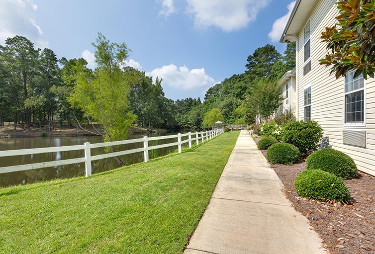 A sidewalk next to a grassy area with a white fence and trees, in a residential neighborhood.