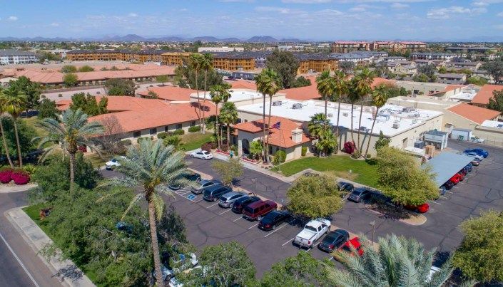 An aerial view of a property with buildings featuring red-tiled roofs, surrounded by palm trees and a parking lot filled with cars.