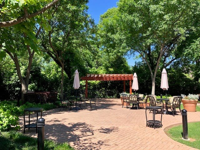 A tranquil garden patio with tables and chairs under the shade of lush trees.