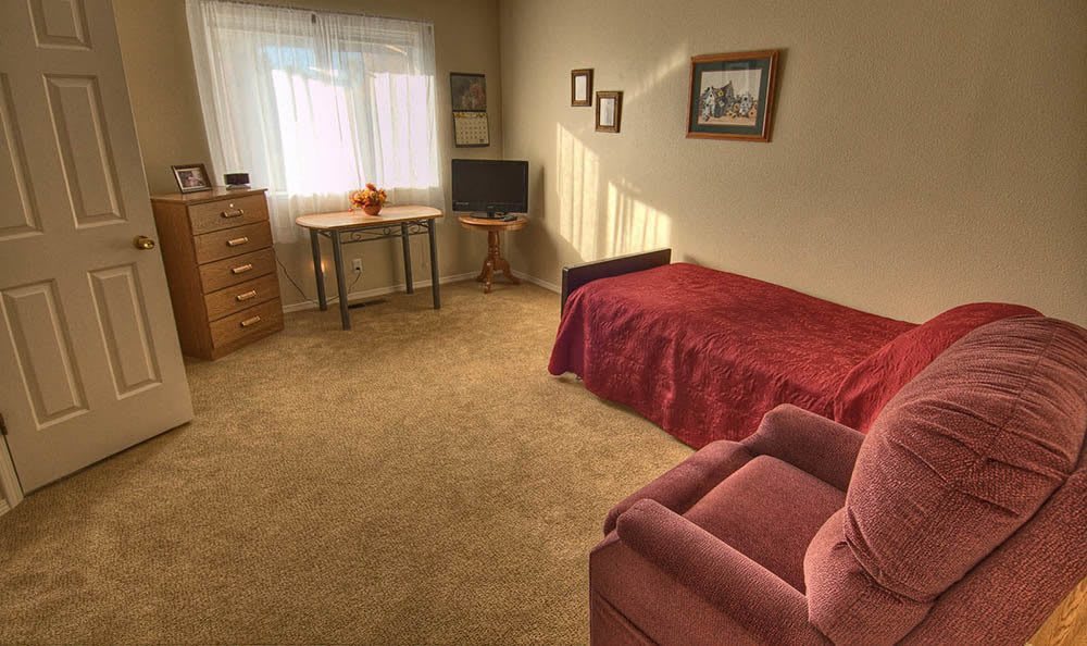 A cozy bedroom with a single bed, armchair, desk, and a window allowing natural light to fill the room.