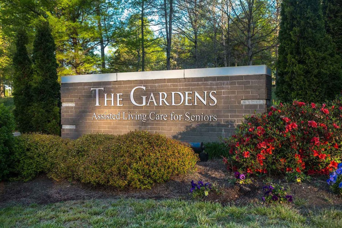 The entrance sign to The Gardens, surrounded by vibrant flowers and lush greenery.