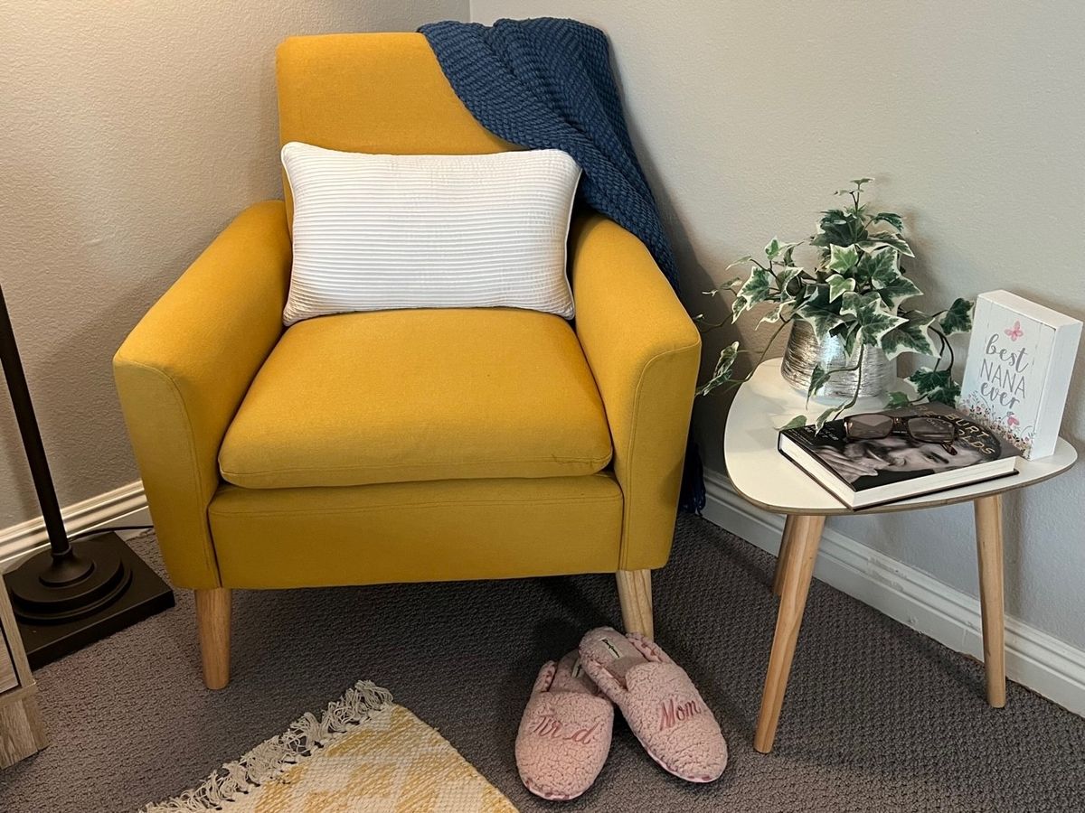 A cozy reading nook with a yellow armchair, draped blanket, and fluffy slippers next to a side table topped with a plant and books.