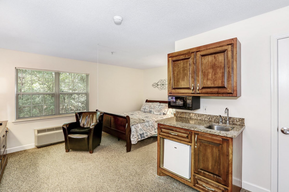 A cozy studio apartment featuring a bed, kitchenette, and seating area with a window view.