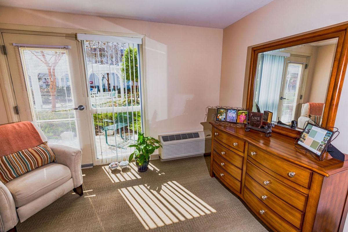 A cozy living room with a chair, a dresser topped with a mirror, and sunlight streaming through a window.