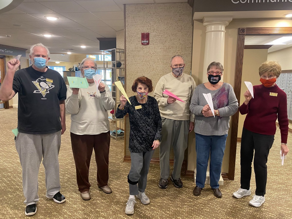 A group of elderly people gather indoors, wearing masks and holding paper airplanes.