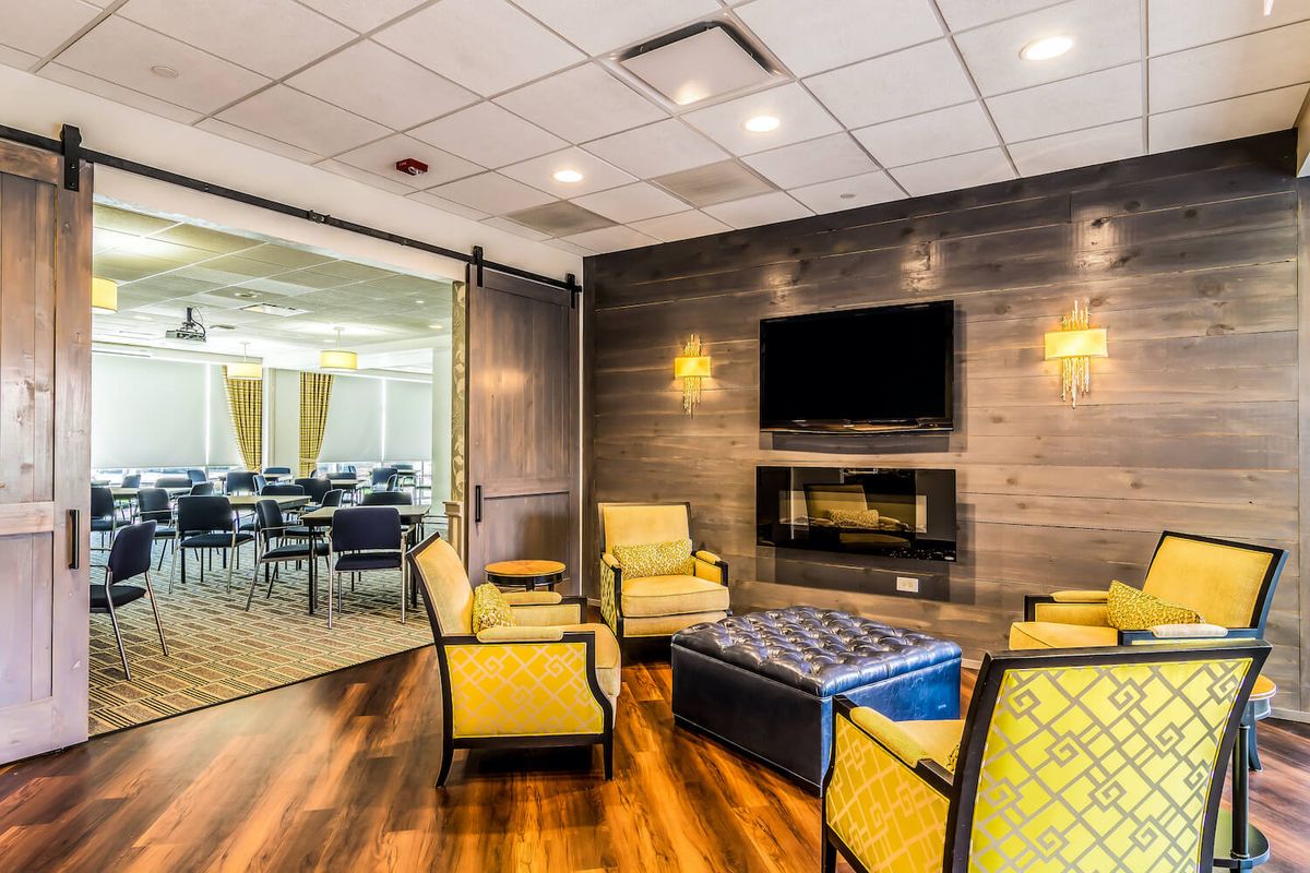 A modern lounge area featuring comfortable seating and a television, with a view into an adjacent meeting room.