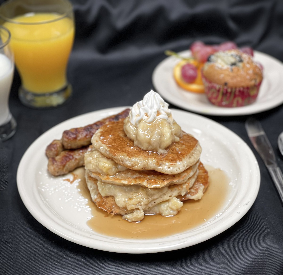 A delicious breakfast featuring pancakes topped with whipped cream and syrup, accompanied by sausages, a muffin, orange juice, and a glass of milk.
