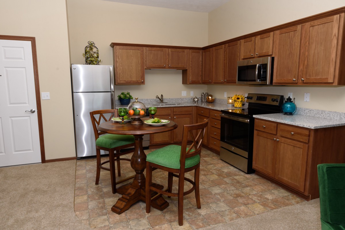 A cozy kitchen with wooden cabinets, stainless steel appliances, and a small dining area featuring a round table with green-cushioned chairs.