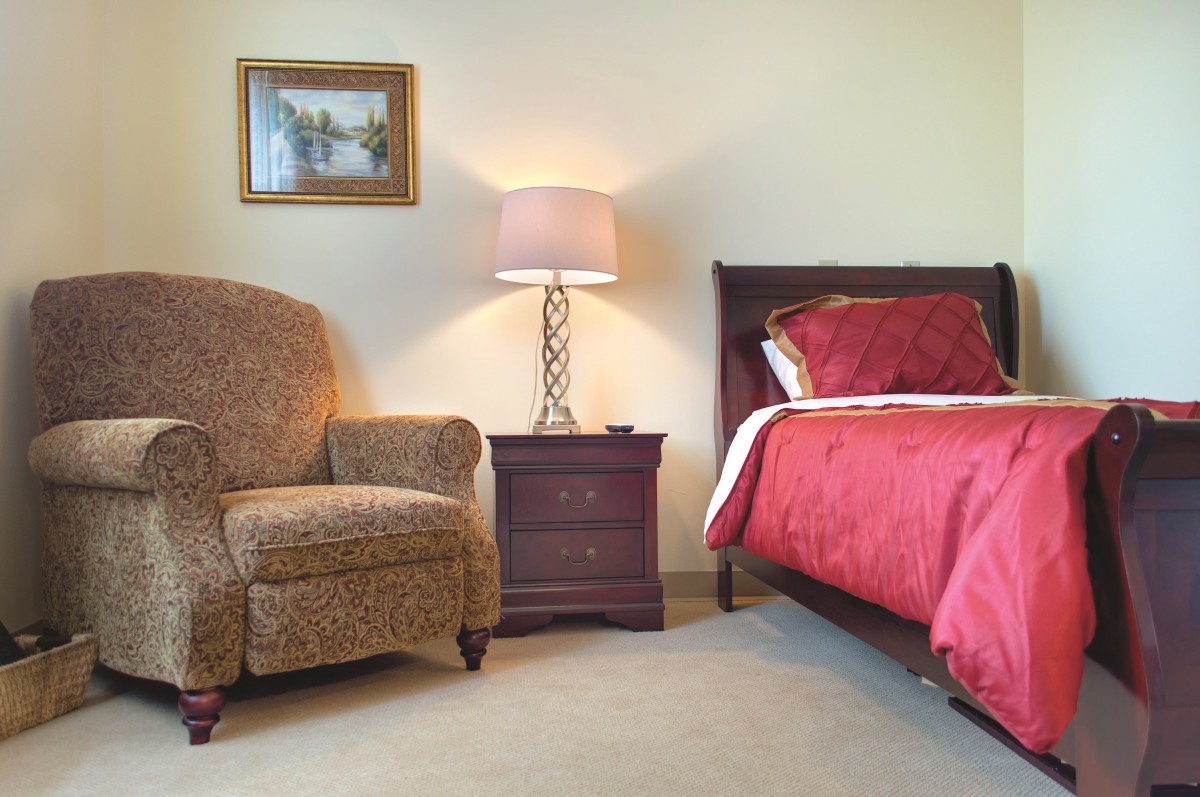 A cozy bedroom featuring an armchair, nightstand with a lamp, and a neatly made bed.