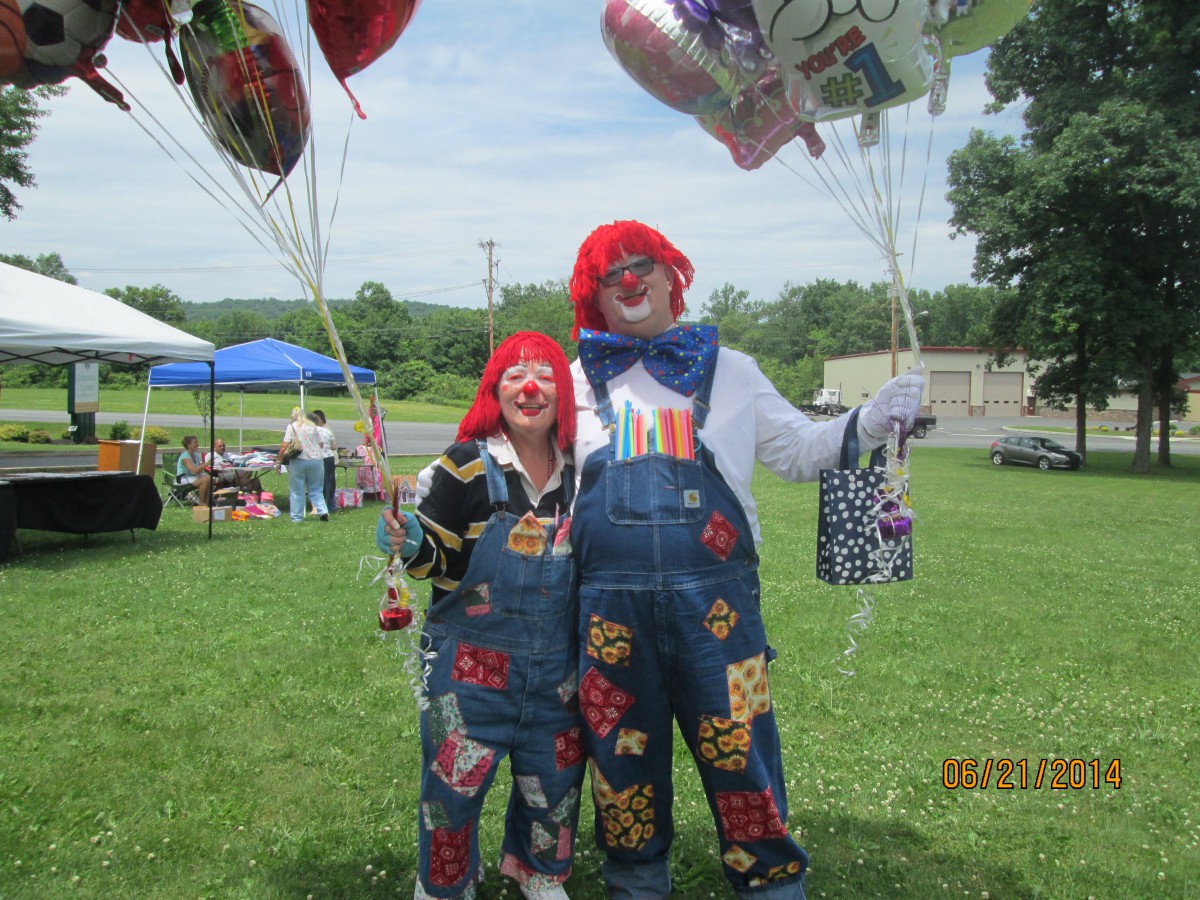 Two clowns in colorful overalls holding balloons at an outdoor event.