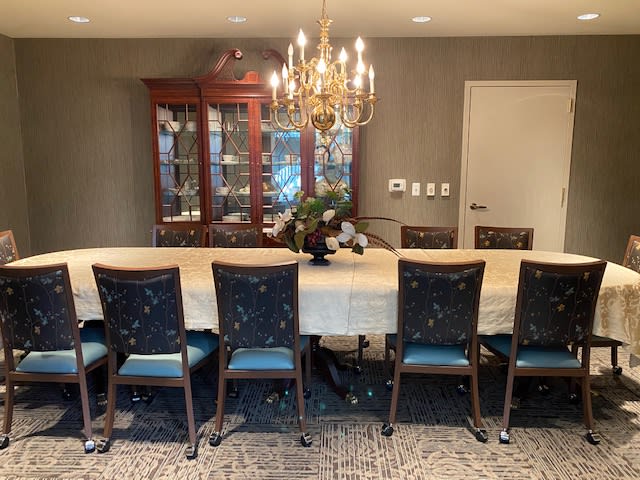 A well-appointed dining room with a long table, surrounded by chairs, and illuminated by a chandelier. A glass cabinet is situated in the background.