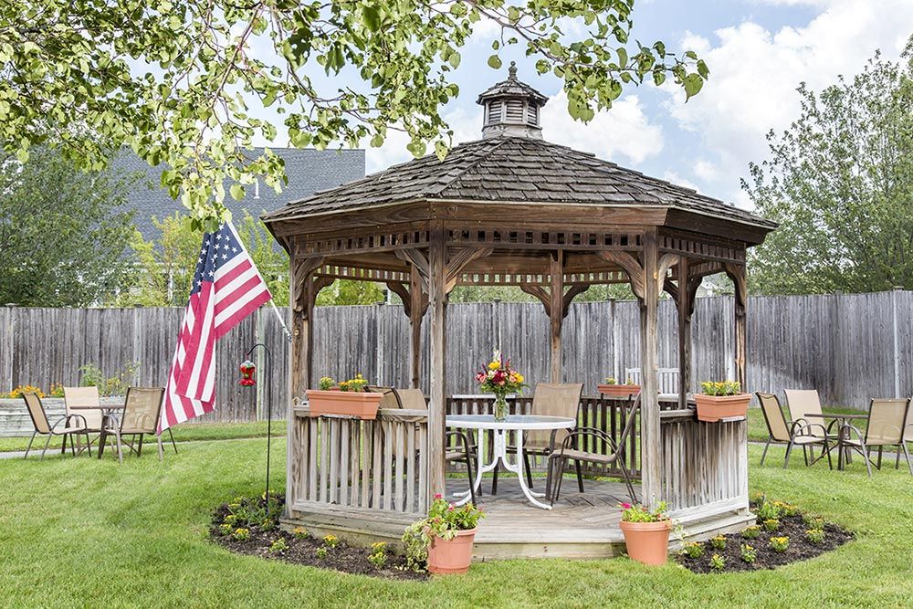 A wooden gazebo in a garden setting with a table and chairs, surrounded by flower pots and an American flag.