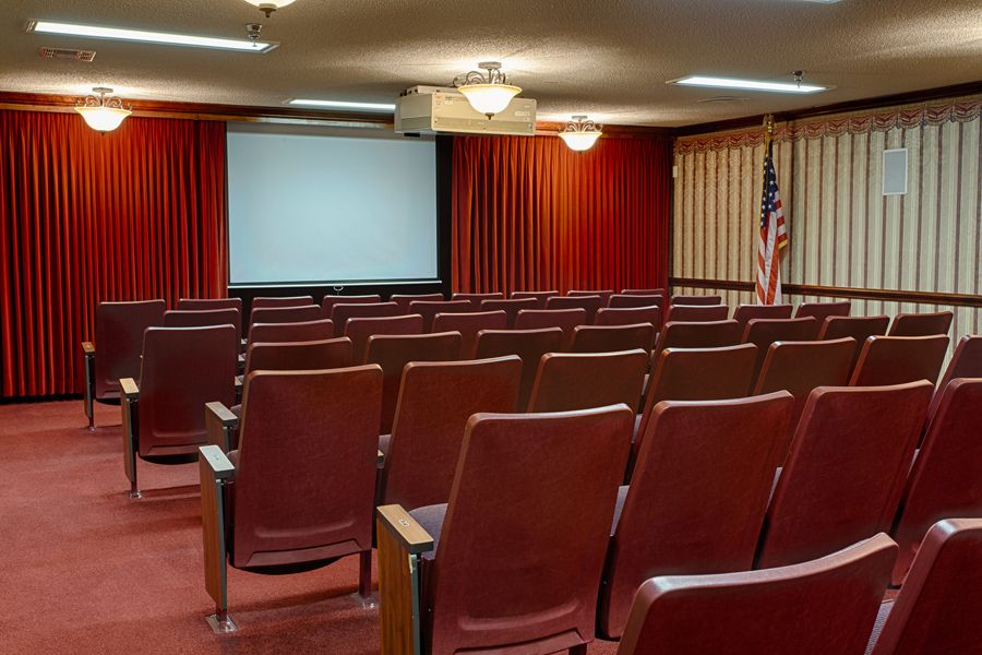An empty auditorium with rows of seats facing a projector screen, featuring red curtains and an American flag.