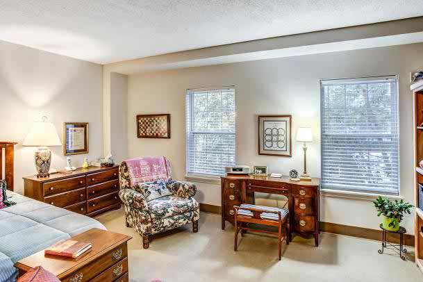 A cozy bedroom featuring classic wooden furniture, including a floral armchair, dressers, and a desk, with natural light streaming through the windows.