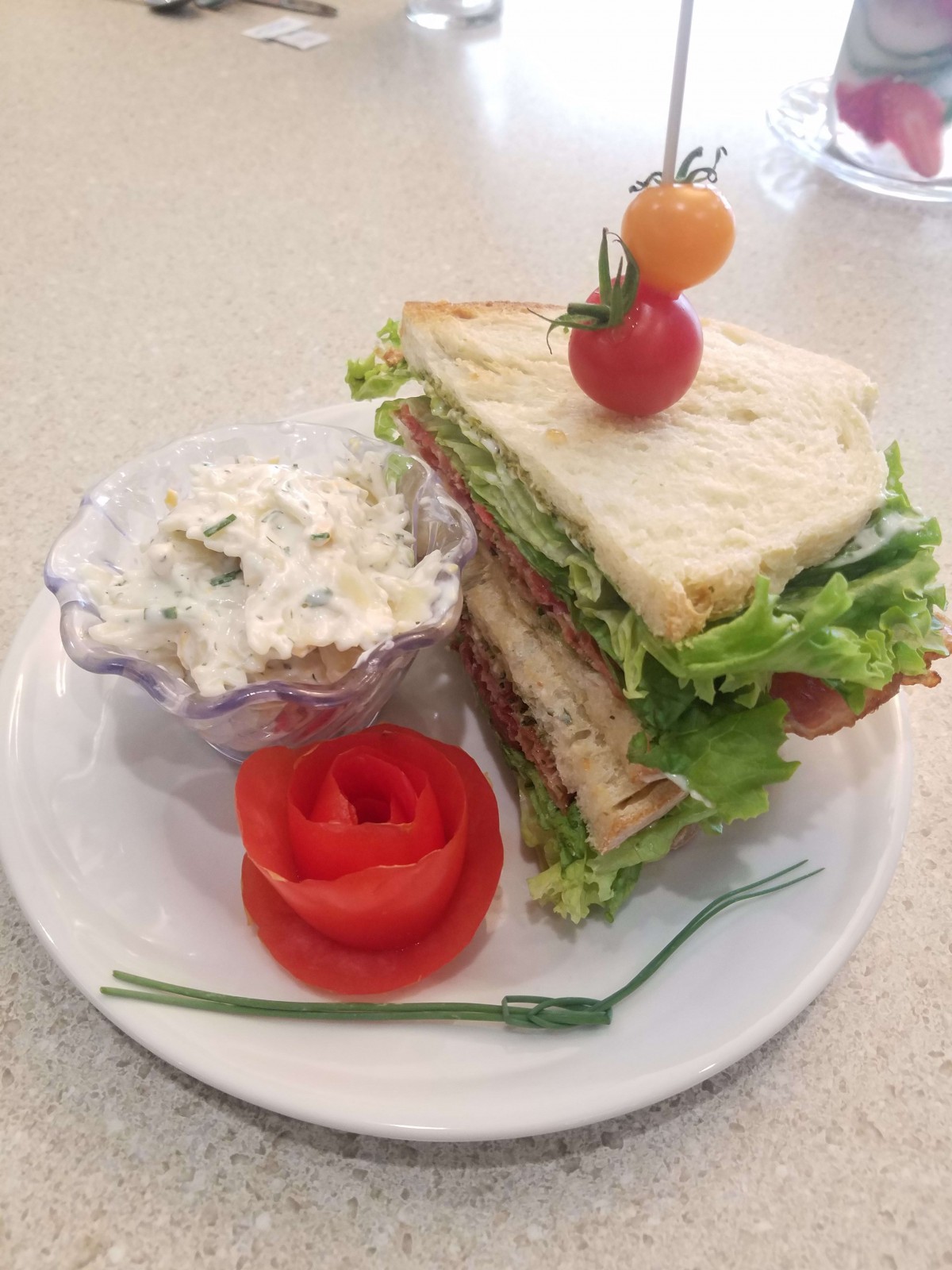 A stacked sandwich with greens and cherry tomatoes, served alongside a creamy salad and garnished with a rose-shaped tomato.