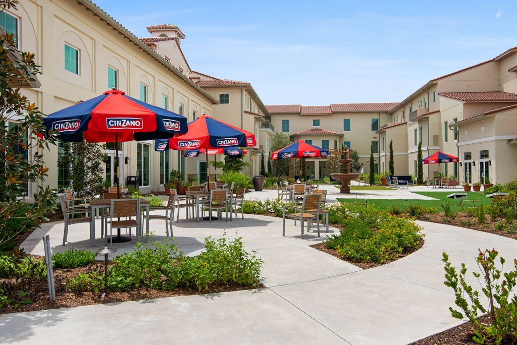 An outdoor patio area with tables, chairs, and umbrellas surrounded by landscaped buildings.